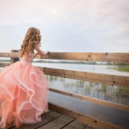 A girl in a flowing pink gown stands on a dock overlooking a serene lake at dusk.